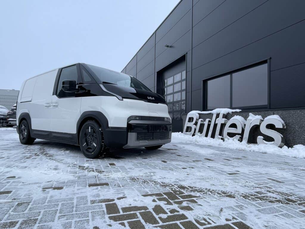 Kleine witte bedrijfswagen voor stadsbezorging met zwart dak en banden, parkeert buiten bij een modern bedrijfsgebouw in winterse omstandigheden.