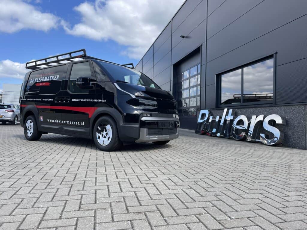 Elektrische Kia PV5 Cargo bedrijfswagen met bedrijfslogo en dakrek, geparkeerd voor een modern bedrijfsgebouw.