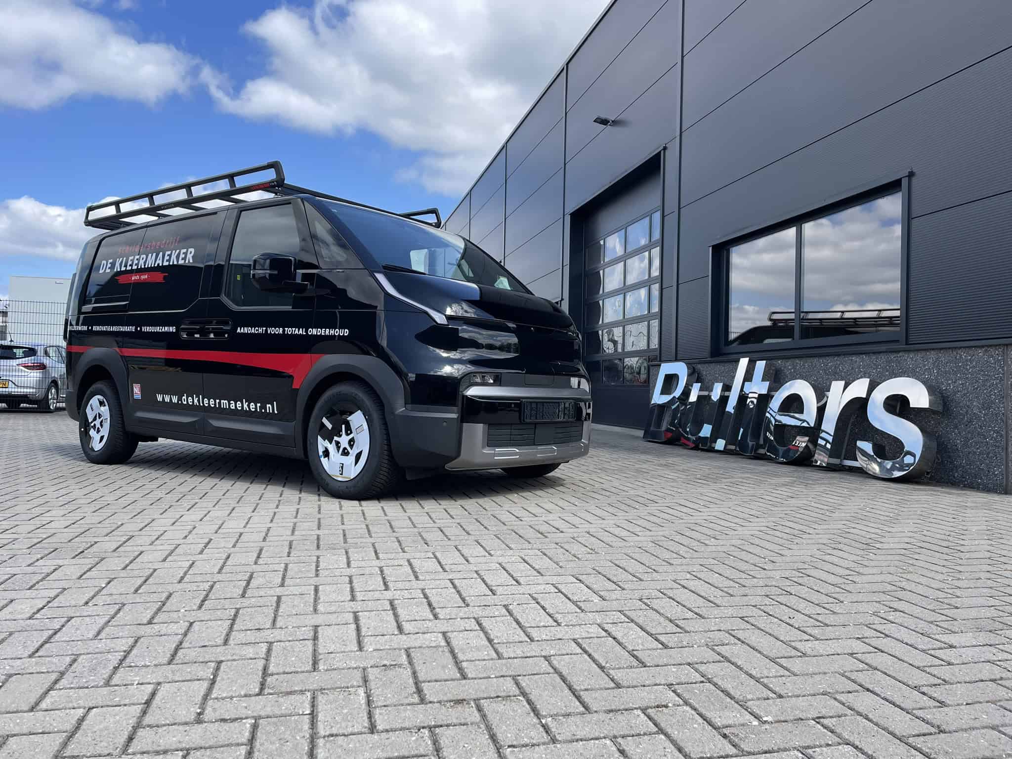 Elektrische Kia PV5 Cargo bedrijfswagen met bedrijfslogo en dakrek, geparkeerd voor een modern bedrijfsgebouw.